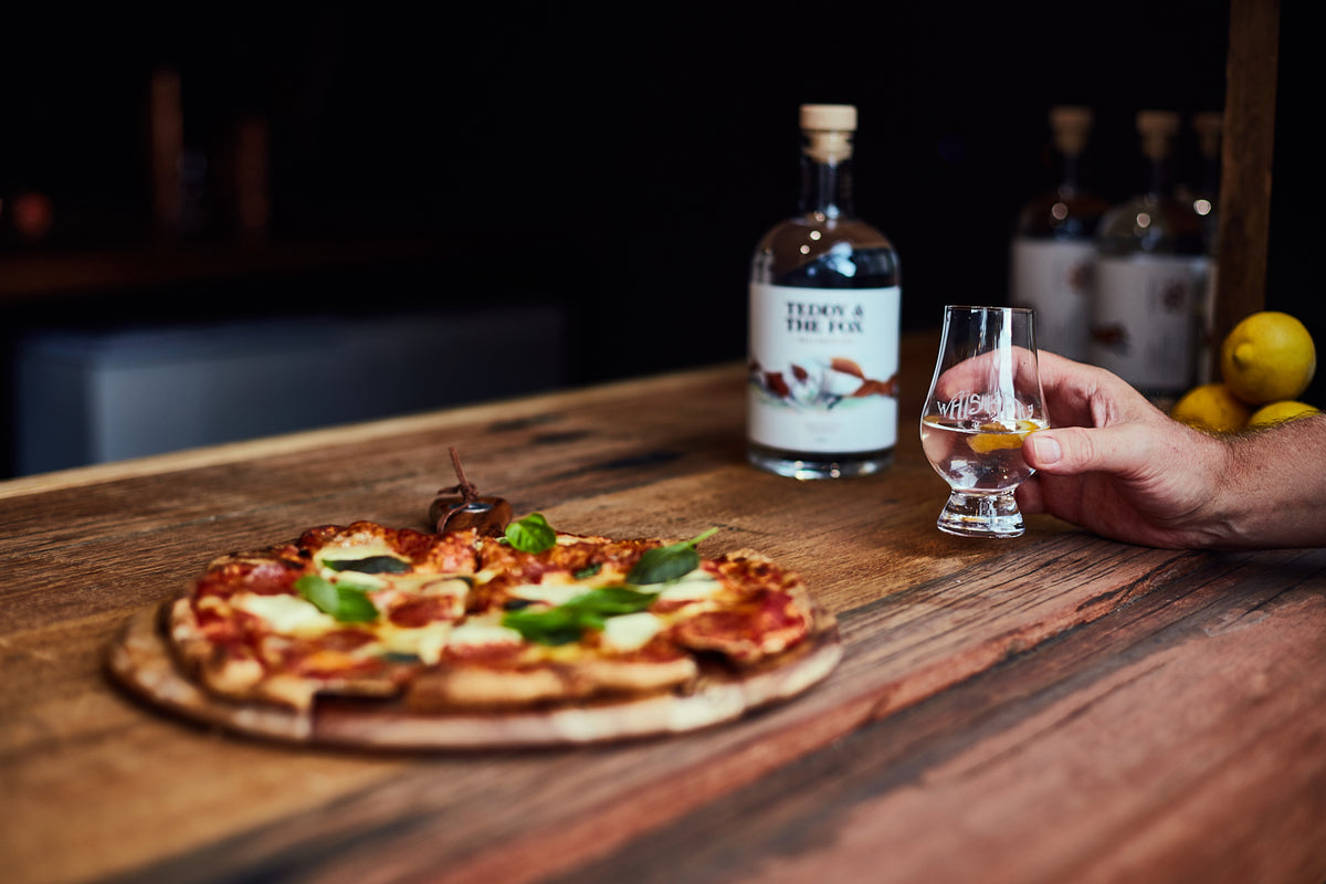 A hand holds a small clear glass with amber liquid beside a branded bottle and a pizza on a wooden table in a dim indoor setting.