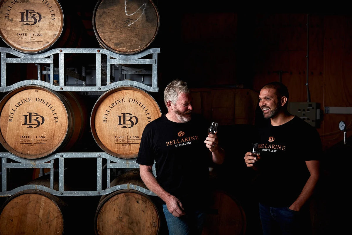 Two men standing in a dimly lit distillery room wearing matching black Bellarine Distillery t-shirts, with stacked wooden barrels in the background.
